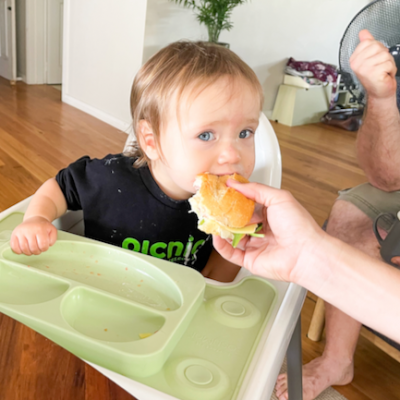 little baby girl in high chair leaning over to take a bite out of her mother sandwich