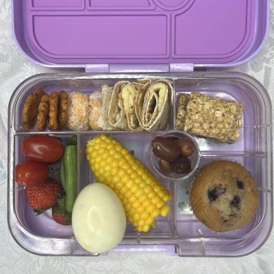 A lunchbox with a variety of foods including a mini muffin, corn on the cob, boiled egg, fruit and vegetables, a wrap, crackers and a muesli bar